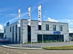 AMPA Kunststof Kozijnen bedrijfspand in Borne. Wit gebouw met blauwe letters. Drie witte vlaggen met het blauwe logo erop op de voorgrond